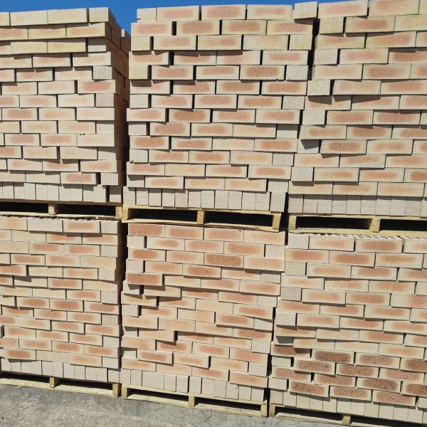 Outdoor yard view of multiple wooden pallets stacked two-high with Letaba Satin face bricks showing pale cream-buff body, light peach-apricot flashed centers, and smooth satin finish.