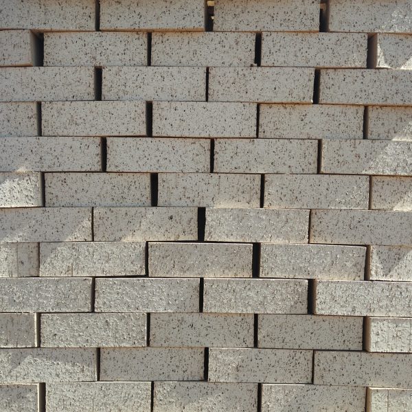 Front-on view of stacked Maize Travertine face bricks showing light cream-maize clay body, pitted travertine texture, and fine manganese iron speckling under sunlight.