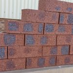 Outdoor pyramid stack of Montana Travertine face bricks displaying pitted travertine texture, rich plum-red clay body, and dark blue-charcoal heat-flashed centers.
