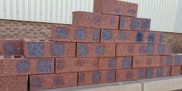 Outdoor pyramid stack of Montana Travertine face bricks displaying pitted travertine texture, rich plum-red clay body, and dark blue-charcoal heat-flashed centers.