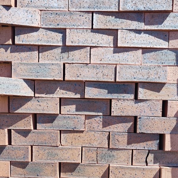 Front-on view of stacked Moroccan Red Travertine Dark face bricks showing pitted travertine surface texture, terracotta-red clay body, and heavy blue-charcoal heat-flashing under direct sunlight.