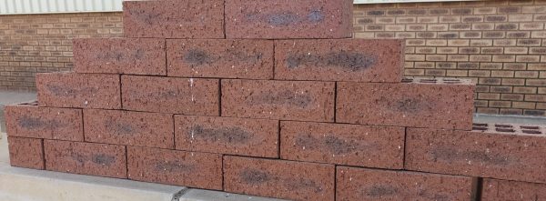 Outdoor pyramid stack of Namib Red Travertine face bricks displaying pitted travertine texture, rich deep red clay body, and dark charcoal heat-flashed centers.