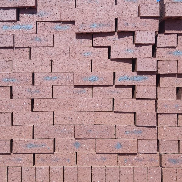 Front-on view of stacked Namib Red Travertine face bricks showing pitted travertine texture, rich deep red clay body, and dark charcoal heat-flashed central markings under direct sunlight.