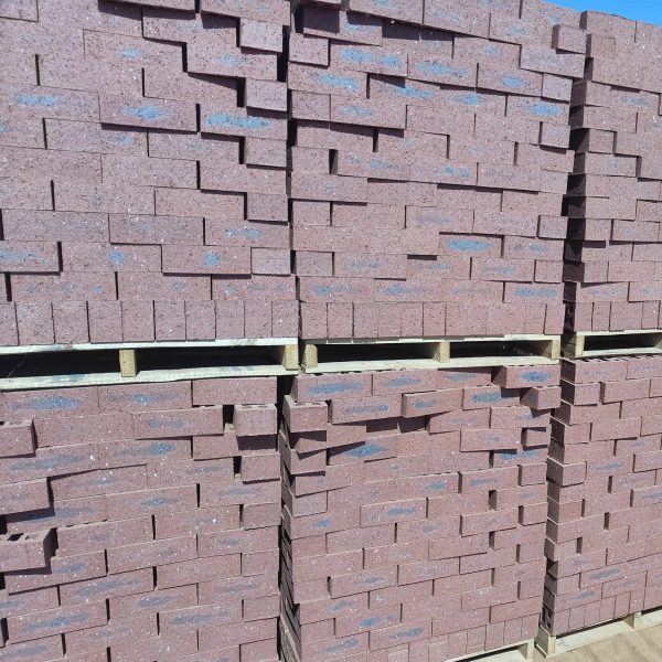Outdoor yard view of multiple wooden pallets stacked two-high with Namib Red Travertine face bricks showing deep red clay body, dark blue-charcoal heat-flashed centers, and pitted travertine finish.