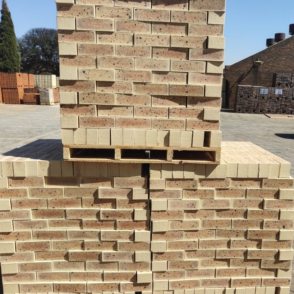 Outdoor factory yard view of Sapphire Satin Light face bricks stacked two-high on wooden pallets under direct sunlight, displaying light cream-buff body, subtle golden flashing, and dark iron speckling.