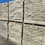 Outdoor factory yard view of Savanna Satin face bricks stacked two-high on wooden pallets under bright blue sky, displaying light wheat-cream body, dark charcoal-purple central flashing, and iron spot speckling.