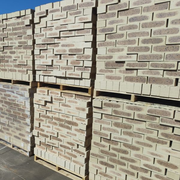 Outdoor factory yard view of Savanna Satin face bricks stacked two-high on wooden pallets under bright blue sky, displaying light wheat-cream body, dark charcoal-purple central flashing, and iron spot speckling.