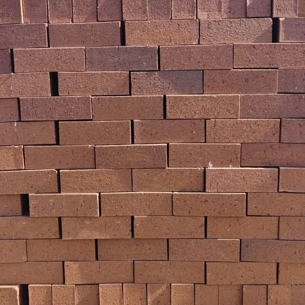 Front-on view of stacked FBA Terracotta Travertine face bricks showing pitted travertine texture, deep terracotta-red clay body, and subtle dark heat-flashing highlights under direct sunlight.