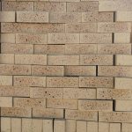 Front-on view of stacked Topaz Travertine face bricks showing pitted travertine surface texture, light golden-buff clay body, warm tan heat-flashed reduction centers, and heavy dark iron speckling.