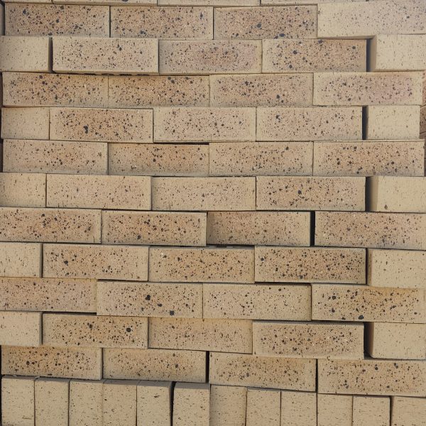 Front-on view of stacked Topaz Travertine face bricks showing pitted travertine surface texture, light golden-buff clay body, warm tan heat-flashed reduction centers, and heavy dark iron speckling.