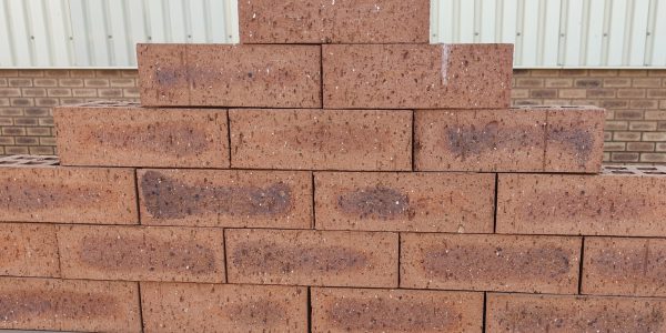 Outdoor pyramid stack arrangement of Wild Wheat Travertine face bricks showing pitted travertine surface texture, earthy red-brown clay body, and dark charcoal heat-flashed centers.
