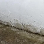 Rising damp and flaking paint where plaster meets paving at the base of a white wall